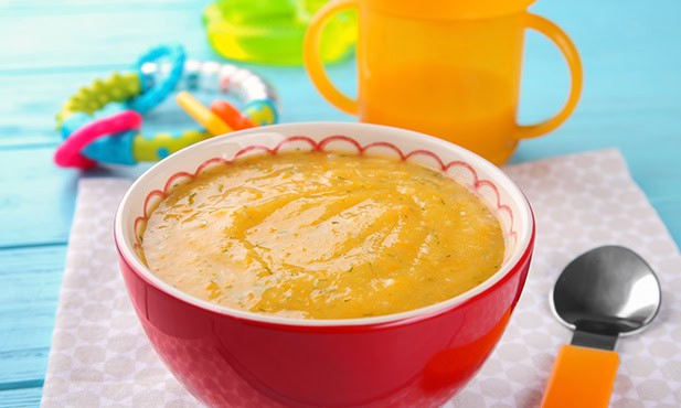 Tigela vermelha com sopa cremosa de legumes para bebê, acompanhada de copo de plástico e colher para bebe ao lado, sobre mesa de madeira clara.